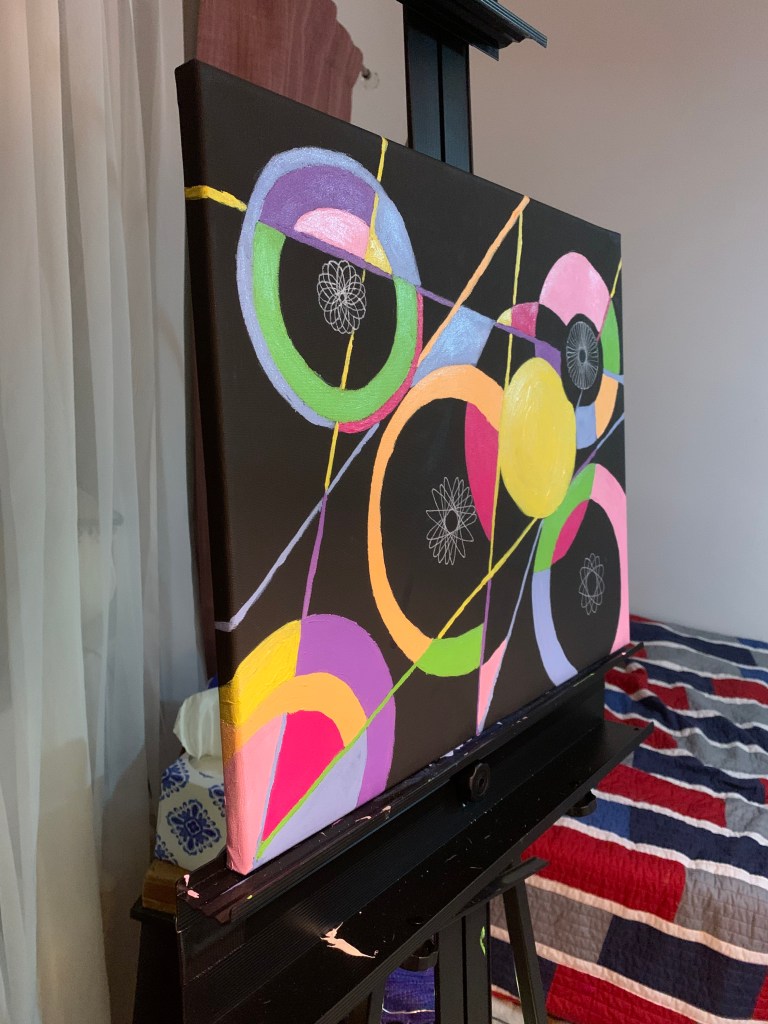 Side view of a black canvas on an easel with pop art circles and lines in pink, purple, green, blue, and yellow, with four white spiral circles created with a spirograph.